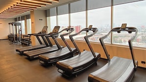 Ein Fitnessraum mit Laufbändern, die an einer Wand entlang verlaufen, mit großen Fenstern und Blick auf die Stadt.