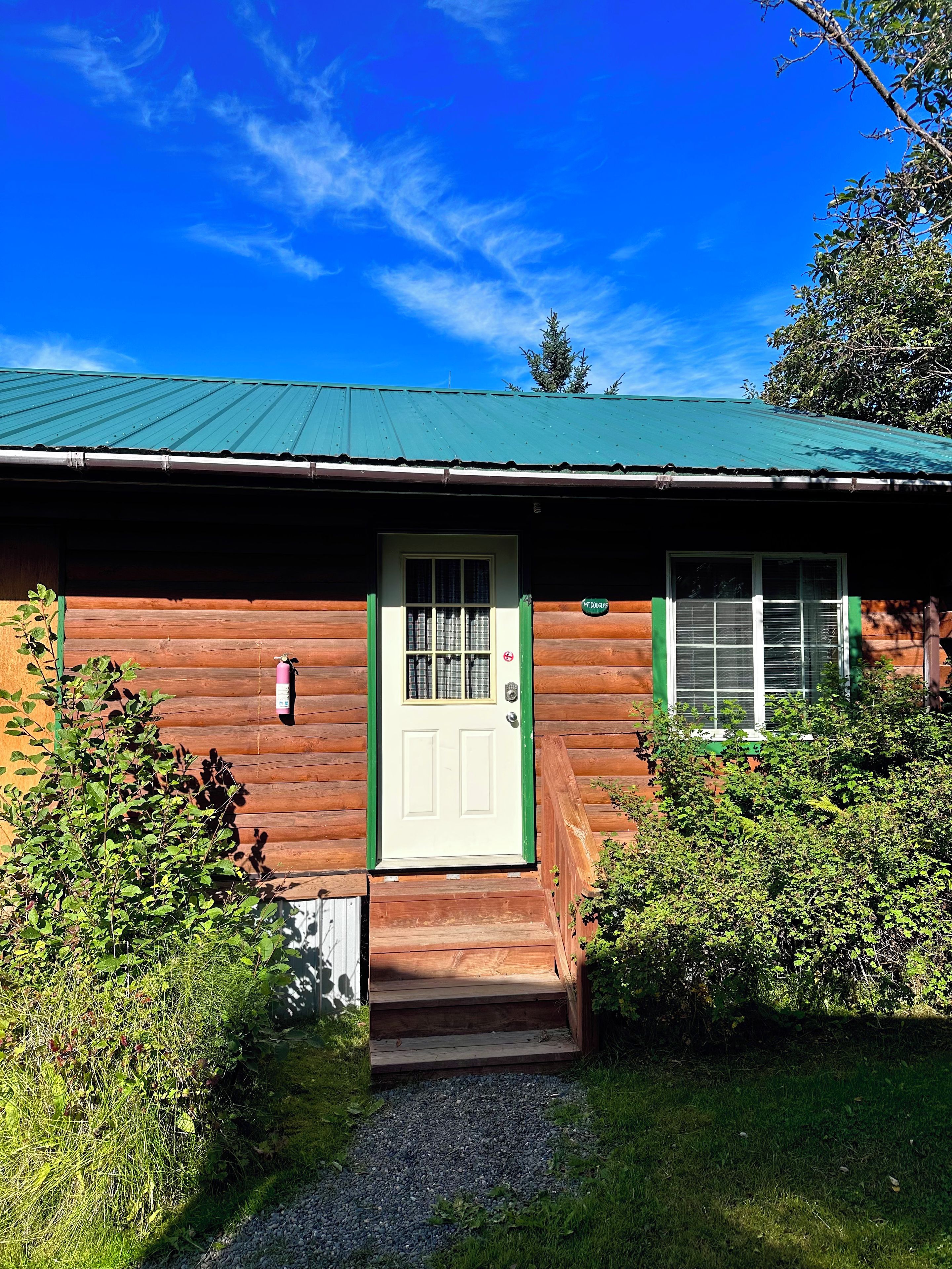 A wooden cabin with a green roof, a white door, and a small porch.