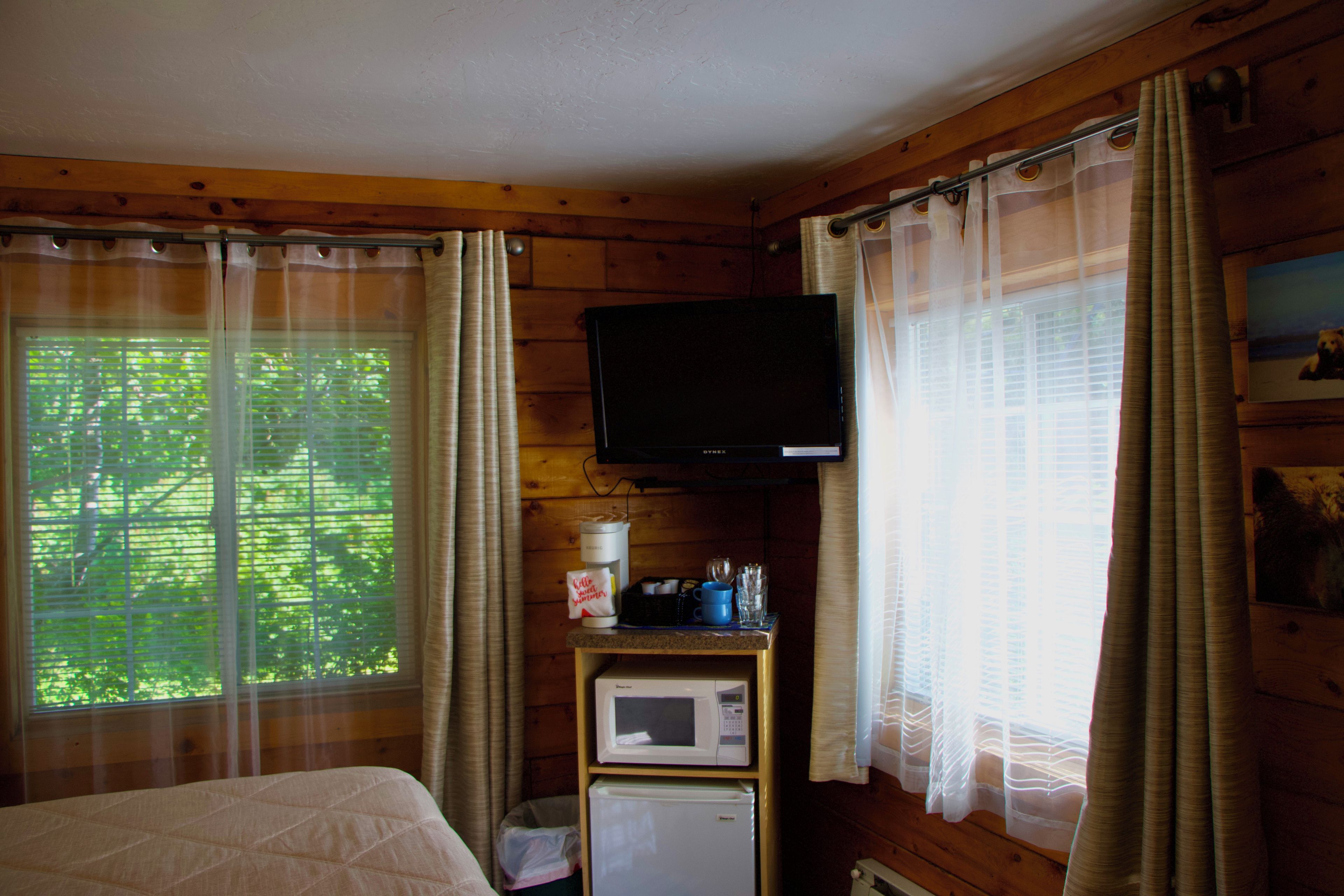A room with a bed, a television mounted on the wall, a small refrigerator, and a window with curtains.
