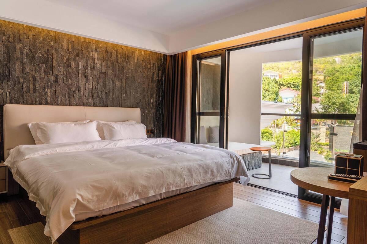 A modern bedroom with a large bed, a small table, and a view of greenery outside.
