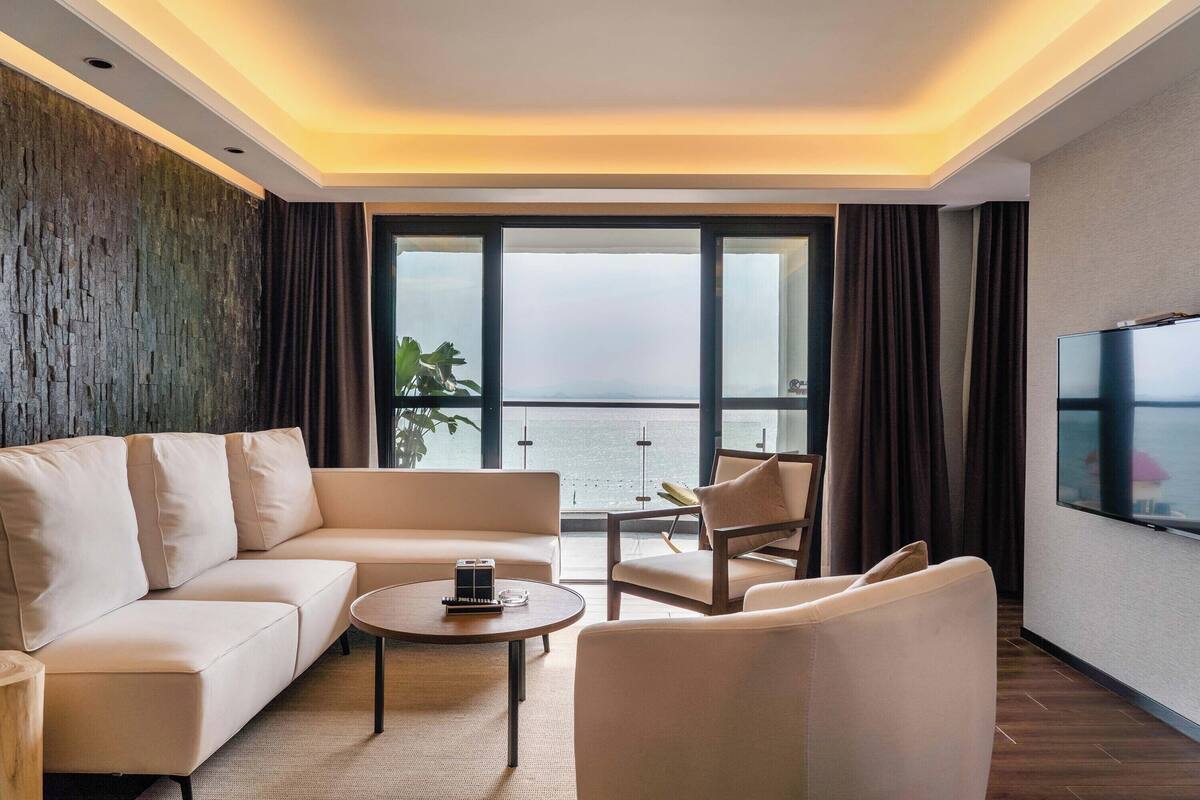 A modern hotel room with a sofa, armchairs, a coffee table, a flat-screen TV, and a large window offering a view of the sea.