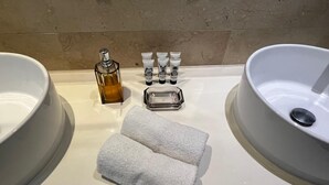 Un baño con dos lavamanos grandes, una botella de jabón líquido, varios tubos de crema, un jabonera de vidrio y dos toallas.