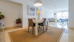 Un comedor moderno con una mesa blanca, sillas e ideal lámpara colgante. Hay dos cuadros en la pared, una planta en maceta y se ve una parte del área de estar al fondo.