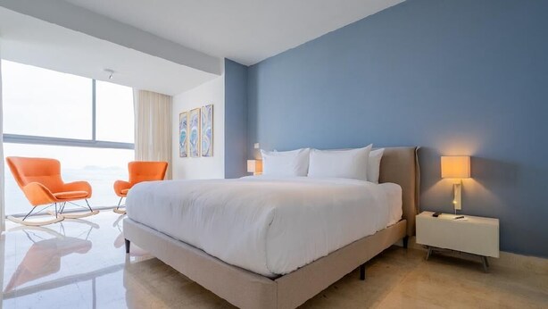 Habitación de hotel moderna con una cama grande, dos sillones naranjas, una mesita de noche y vistas de la ciudad.