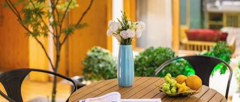 Una mesa de madera redonda con un periódico, un florero con flores blancas y una cesta de frutas.