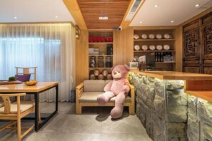 A cozy room with a wooden dining table, a cushioned chair, a large teddy bear, and a stone accent wall. - UNThinkHotel Qimu inn (Lijiang)