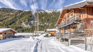 Una cabaña de madera con balcón, rodeada de nieve y montañas al fondo.