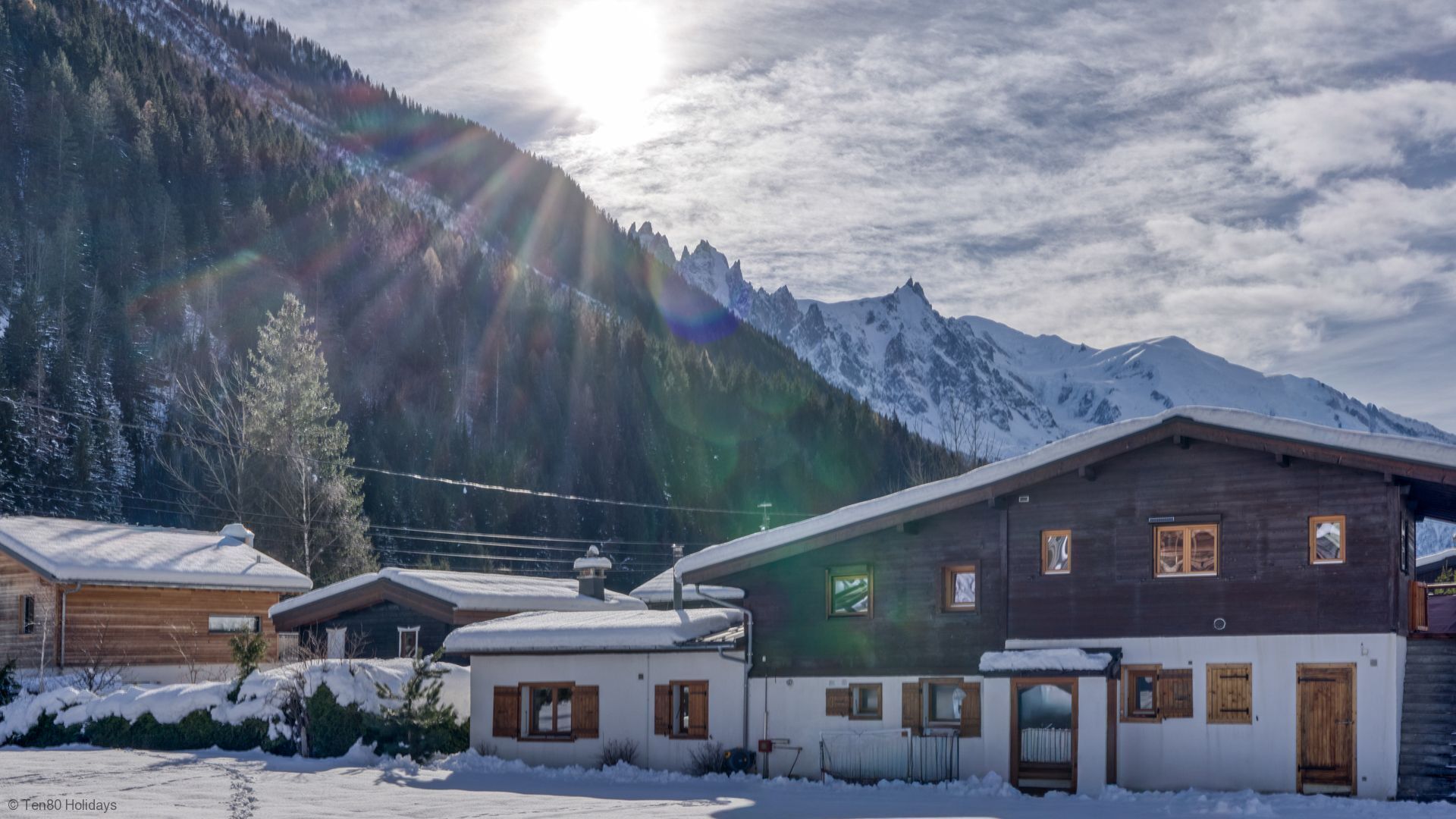 Une cabane en bois dont le toit est recouvert de neige, entourée d’un paysage enneigé et avec des montagnes en arrière-plan.