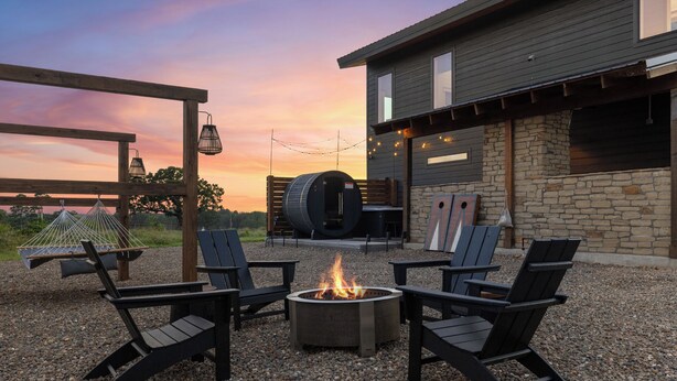 An outdoor seating area with four Adirondack chairs around a fire pit, a wooden pergola, and a stone wall with a barrel and a wooden bench. - Ranch & Relax - Sauna, Hot Tub + Stargazing Escape (Broken Bow)