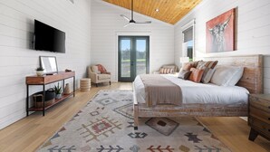 A bedroom with a wooden ceiling, a bed with multiple pillows, a TV, a desk with a plant, and a door leading to a balcony. - Ranch & Relax - Sauna, Hot Tub + Stargazing Escape (Broken Bow)