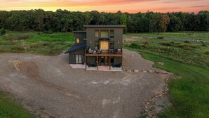 A modern two-story house with a deck, surrounded by a gravel driveway and a grassy field. - Ranch & Relax - Sauna, Hot Tub + Stargazing Escape (Broken Bow)