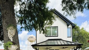 A two-story house with a covered patio, a water tower in the background, and a tree with a sculpture. - The Mini Mansion on Main Street (La Porte)