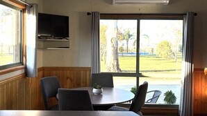 Una habitación con una mesa redonda, sillas, un televisor instalado en la pared y un gran ventanal con cortinas.
