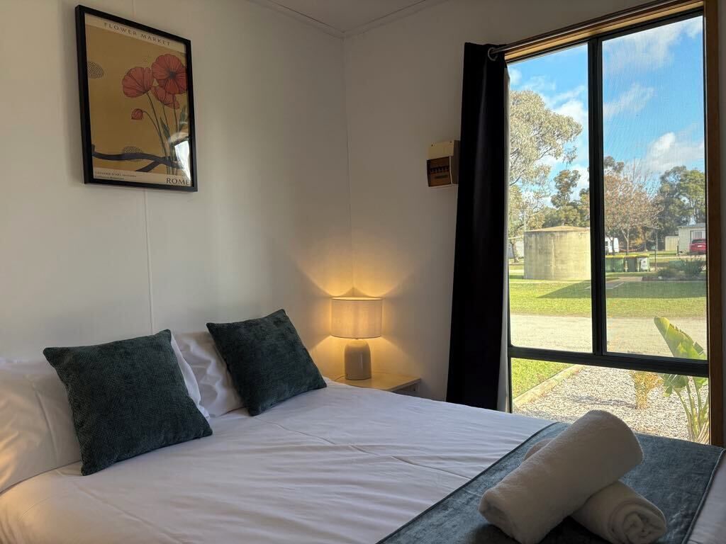 A hotel room with a bed, pillows, a lamp, a framed picture on the wall, and a window with a view of the outdoors.