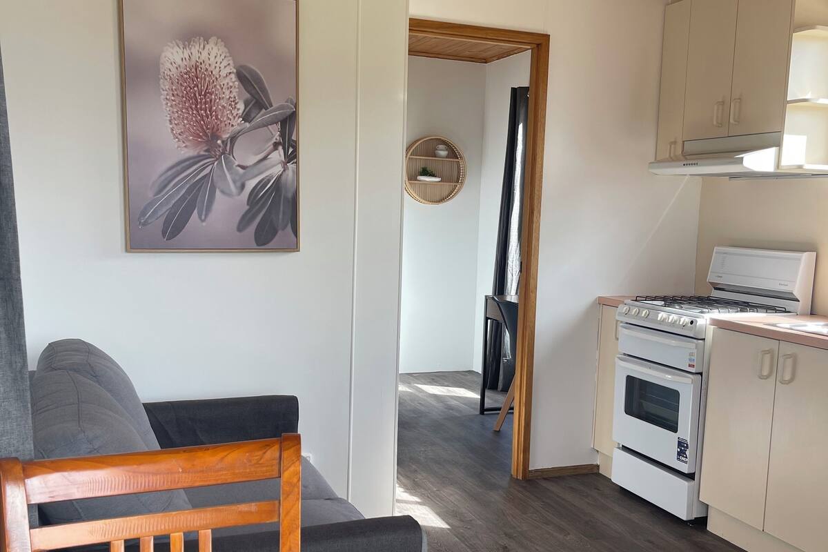 Ett modernt kök med vita skåp, en inbyggd ugn och en spishäll. Det finns en matplats med ett träbord och en grå soffa. En inramad bild av en blomma hänger på väggen.