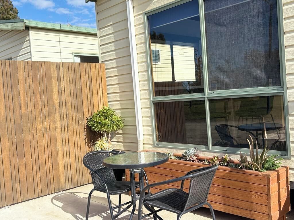 An outdoor patio with a round table and two chairs, a wooden planter with plants, and a large window with a view of a house.