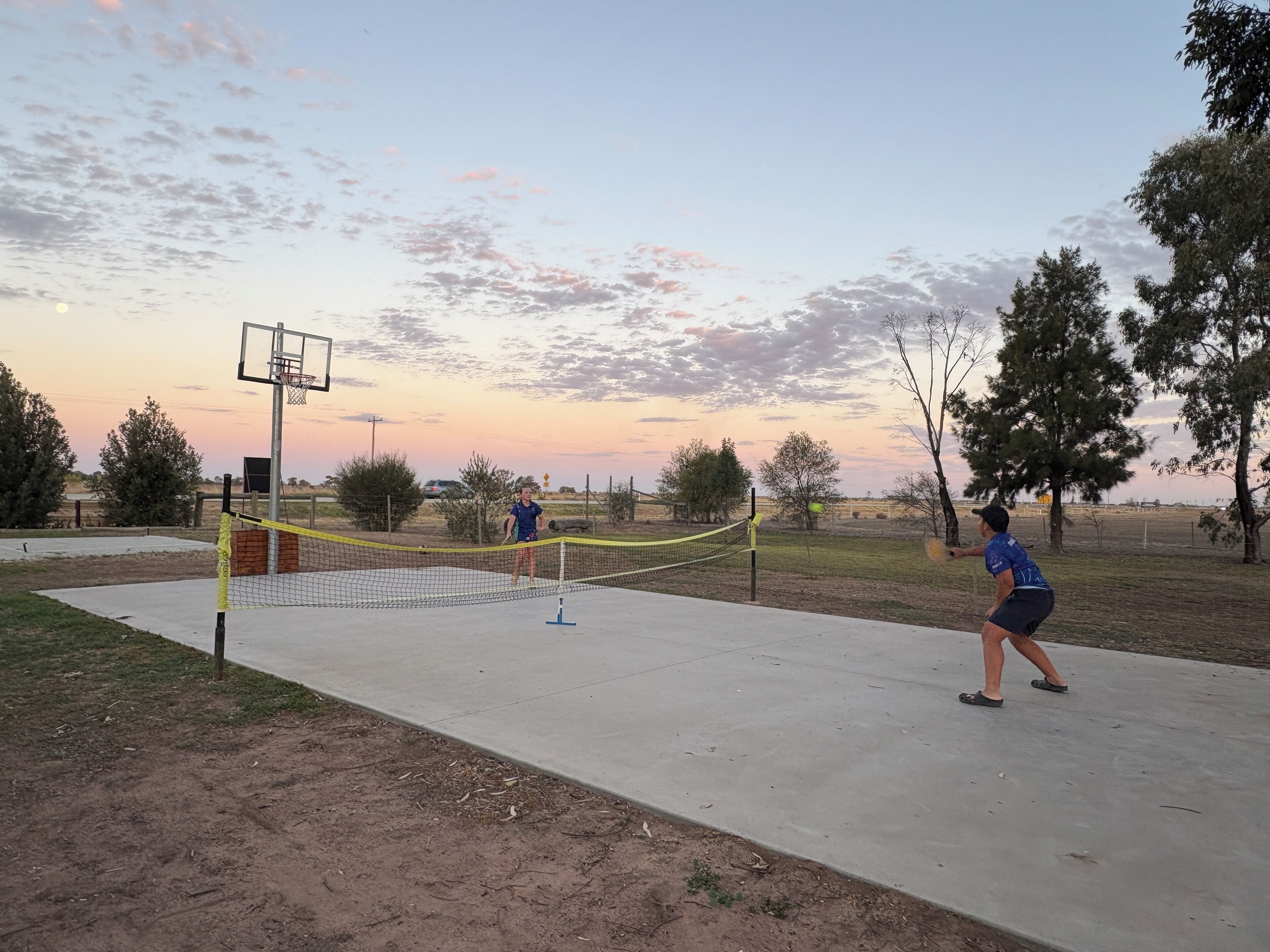 Una cancha de netball al aire libre con un aro de baloncesto y árboles de fondo.