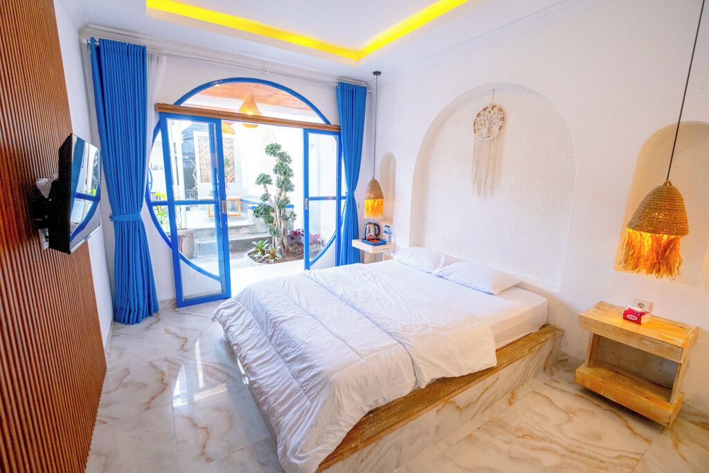 Une chambre avec un lit, une table de chevet en bois, un attrape-rêves accroché au mur et une vue à travers une porte vitrée.