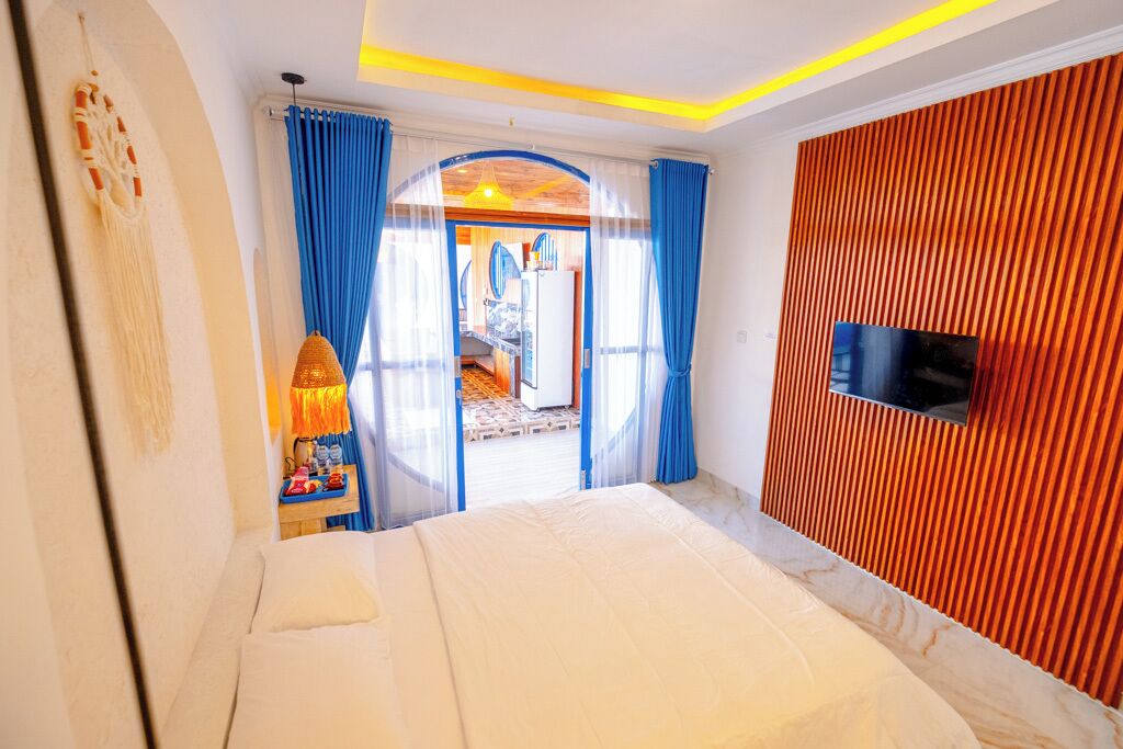 A modern hotel room with a bed, a TV mounted on a wooden panel wall, and a view of the outdoor area through a glass door.