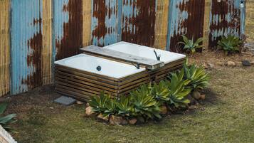 A rectangular outdoor hot tub with a wooden frame, surrounded by rocks and plants, against a rusted metal fence.