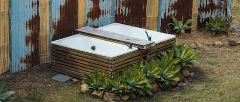 Ein rechteckiges, Outdoor-Whirlpool mit Holzrahmen, umgeben von Steinen und Pflanzen, vor einem rostigen Metallzaun.