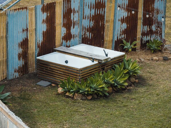 A rectangular outdoor hot tub with a wooden frame, surrounded by rocks and plants, against a rusted metal fence.