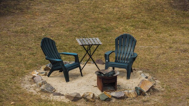 Deux fauteuils Adirondack et une petite table en bois sont disposés sur une pelouse, avec un foyer au centre.
