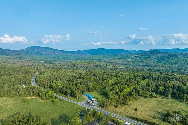 An aerial view of a rural area with a road, a blue building, and surrounding forested landscape. - Cascade Inn (Lake Placid)