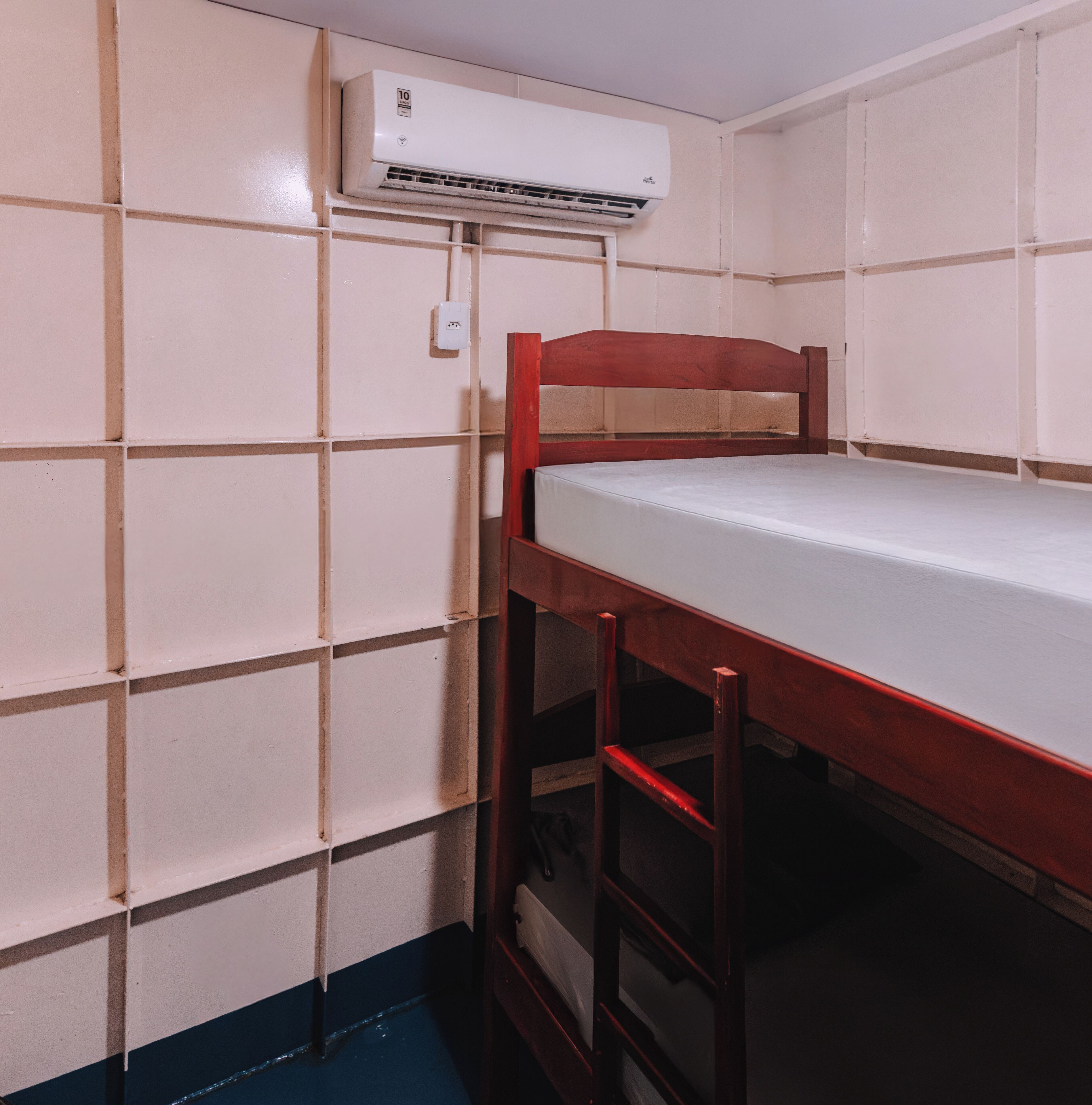 Una cama litera con estructura de madera y colchón blanco, un equipo de aire acondicionado blanco montado en la pared, y una pared de azulejos blancos con sección inferior azul.