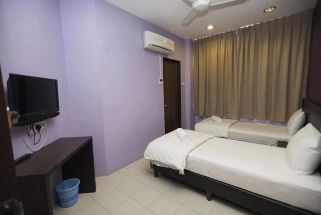 A hotel room with two single beds, a TV mounted on the wall, an air conditioning unit, and a curtain on the window.