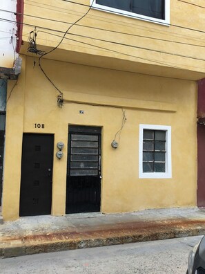 A yellow building with a black door labeled "108," a black gate, and a small window. - Central Accommodation We invoice. (Villahermosa)