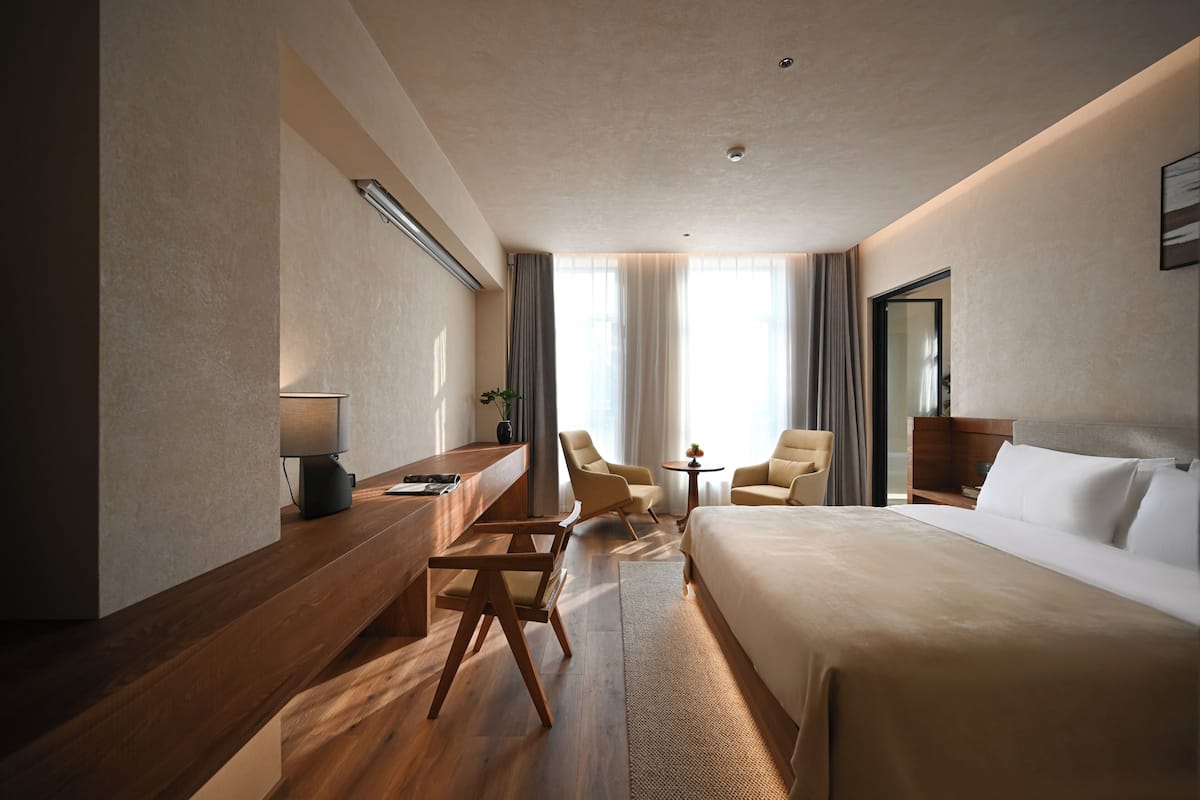A hotel room with a large bed, a desk, two chairs, a small table, and a window with curtains.