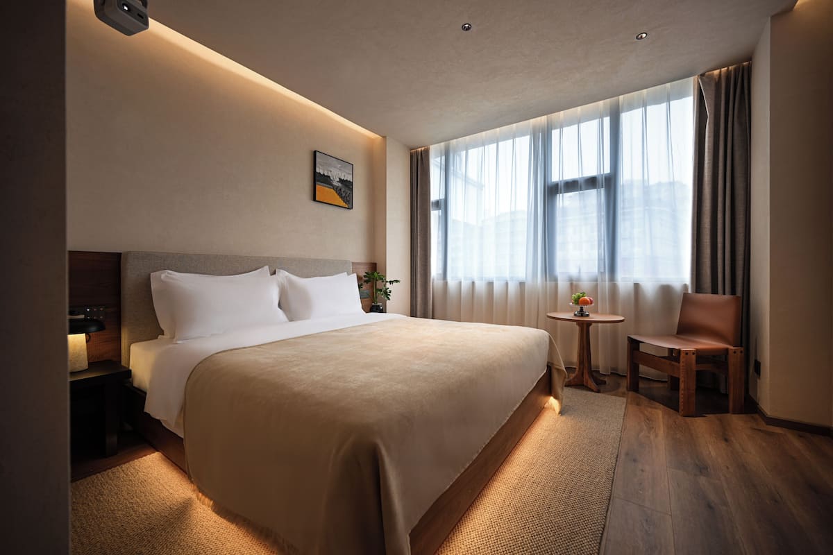A hotel room with a large bed, a small table with a plant, a chair, and a window with curtains.