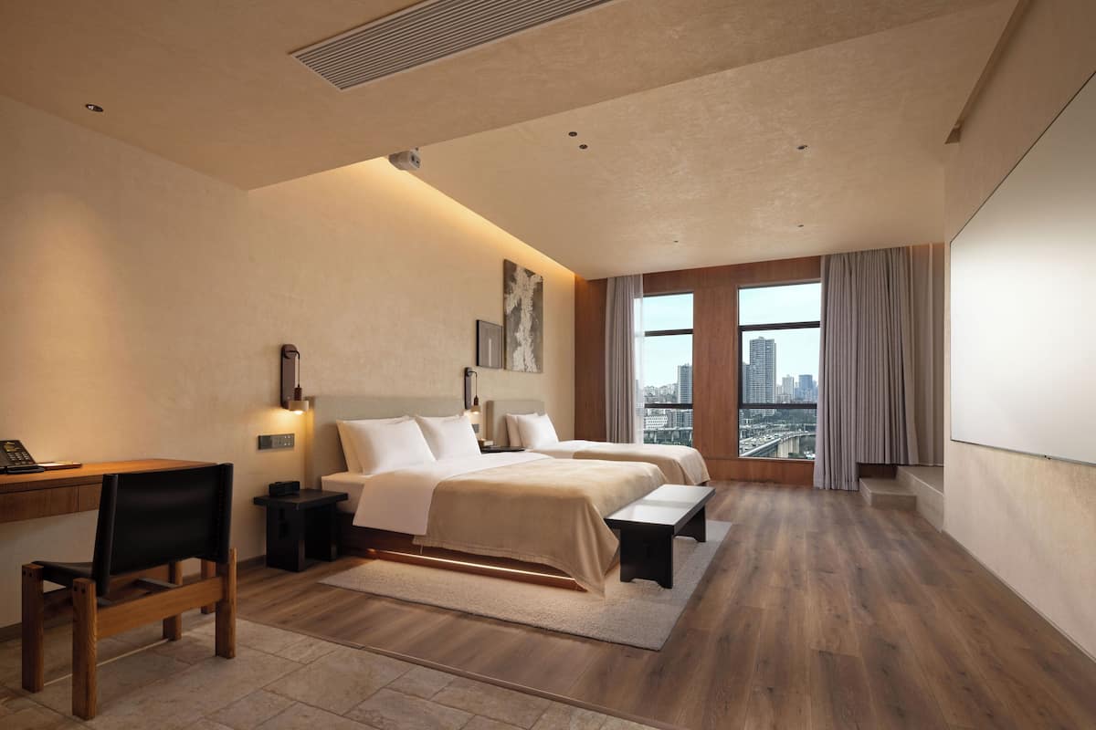 A modern hotel room with a large bed, a desk, a chair, a bench, and a view of the city through large windows.