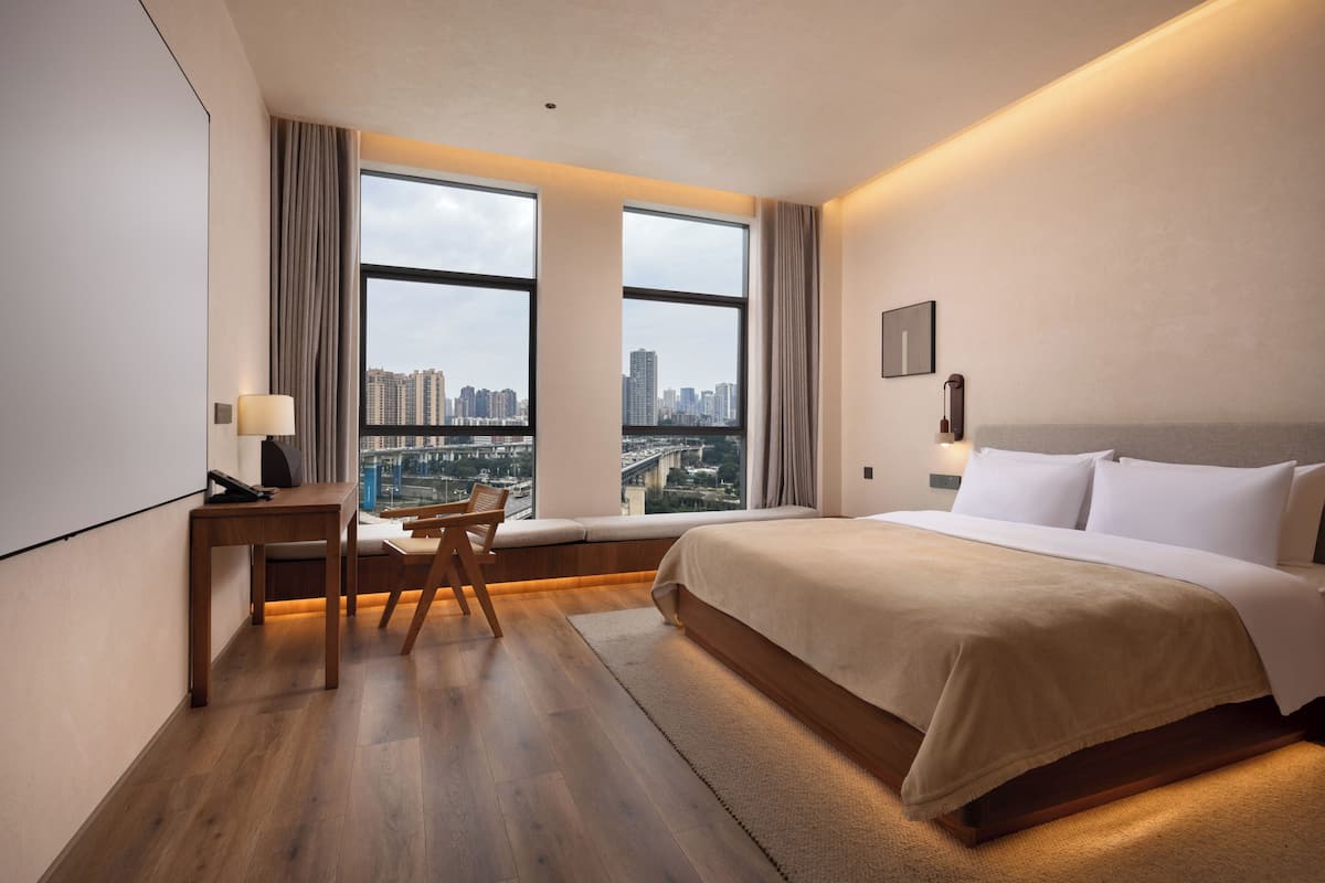 A hotel room with a large bed, a desk, a chair, and a view of the cityscape through large windows.