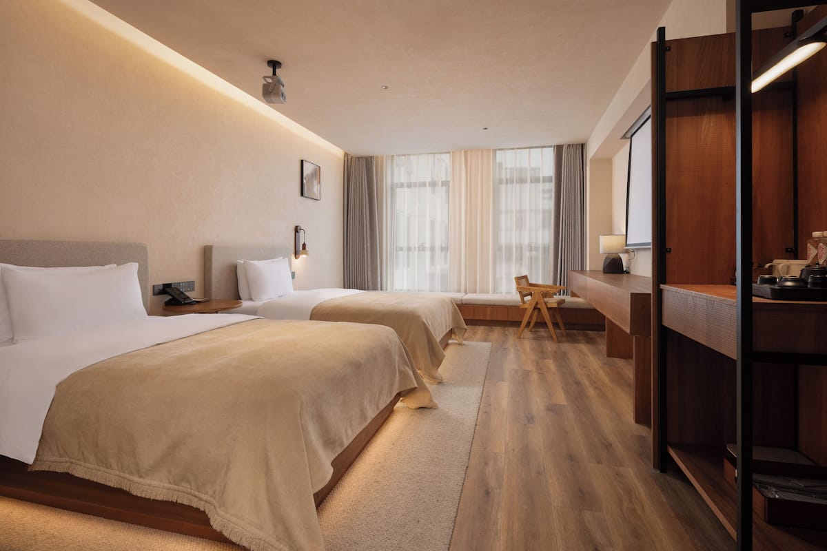 A hotel room with two beds, a desk, a chair, a TV, and a large window.