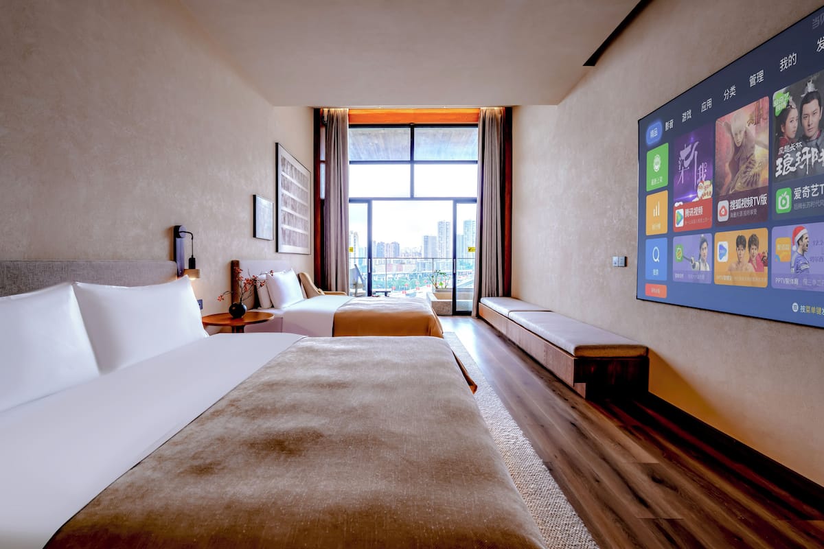 A hotel room with a large bed, a smaller bed, a seating area, and a wall-mounted TV displaying various channels.