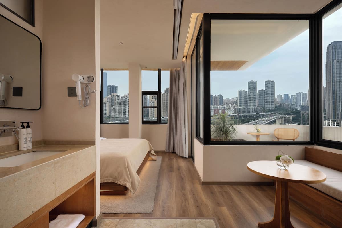 A modern hotel room with a large window, a bed, a bathroom with a vanity, and a small table with a vase.