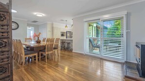 Ruime eethoek met een houten tafel en stoelen, een keuken op de achtergrond en een grote schuifdeur met zonwering.