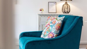Una cadira color aguamarina amb un coixí floral, un certificat emmarcat, un vas petit amb flors i una làmpada sobre una alçada blanca.