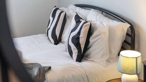 A neatly made bed with white bedding and striped pillows, a bedside lamp, and a wooden nightstand. - No15 Ellis House - Donnini Apartments (Kilmarnock)