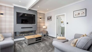 A modern living room with a flat-screen TV mounted on a gray wall, a wooden coffee table, two gray sofas, and a small side table with a lamp. - No15 Ellis House - Donnini Apartments (Kilmarnock)
