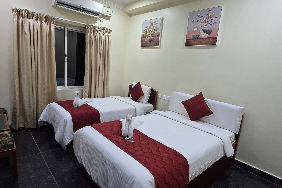 Quarto de hotel com duas camas, janela com cortinas, uma pequena mesa e uma garrafa de água, além de duas pinturas penduradas na parede.
