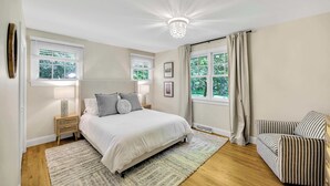 Un dormitorio con una cama grande, dos mesitas de noche con lámparas, un sillón a rayas y vistas a zonas verdes por las ventanas.
