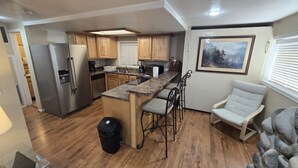Cocina compacta con armarios de madera, refrigerador de acero inoxidable, encimera de granito y una pequeña isla con taburetes de bar.
