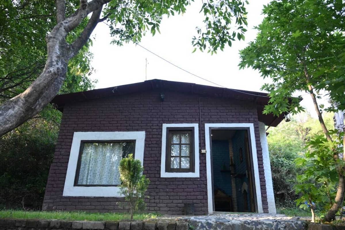 Un pequeño edificio de ladrillo con dos ventanas y una puerta, rodeado de árboles.