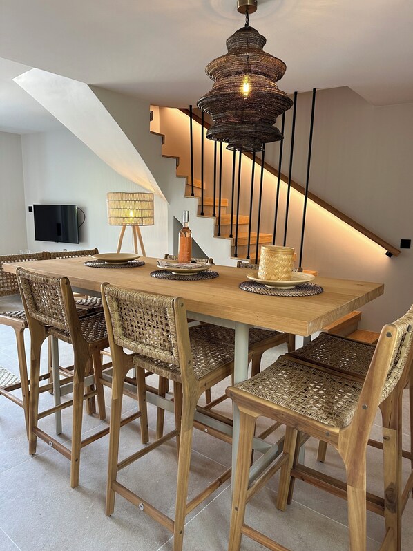 A dining area with a wooden table and wicker chairs, a hanging light fixture, and a staircase with wooden steps and black railings.