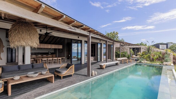 Un área de piscina moderna al aire libre con mobiliario de madera, techo de paja y vista al océano.