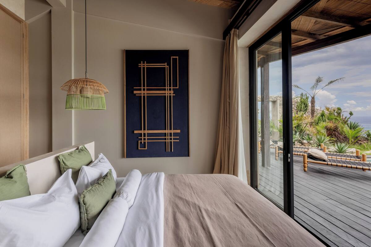 A modern bedroom with a large bed, a hanging light fixture, and a view of a deck with plants.