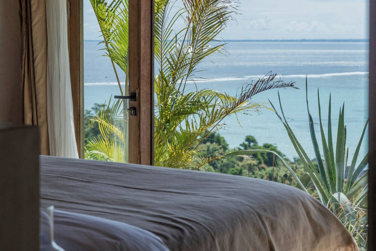 A bedroom with a large window overlooking a beach and ocean.